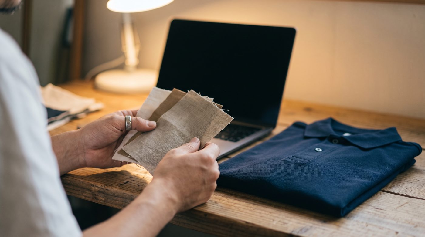 A business owner at a desk comparing plain fabric swatches and a folded plain navy polo shirt beside an open laptop