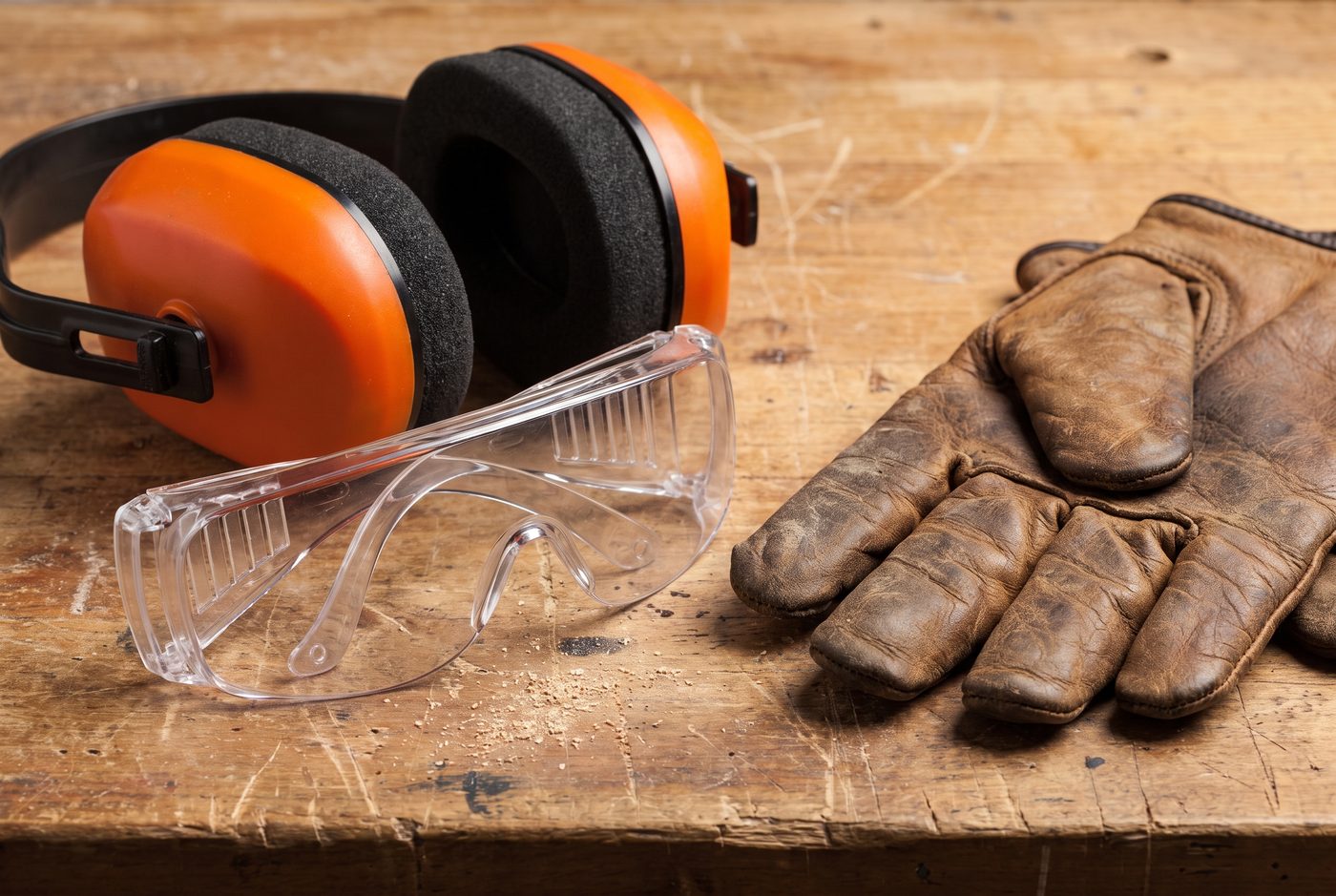 A pair of clear safety glasses and a set of ear defenders laid on a workbench next to work gloves